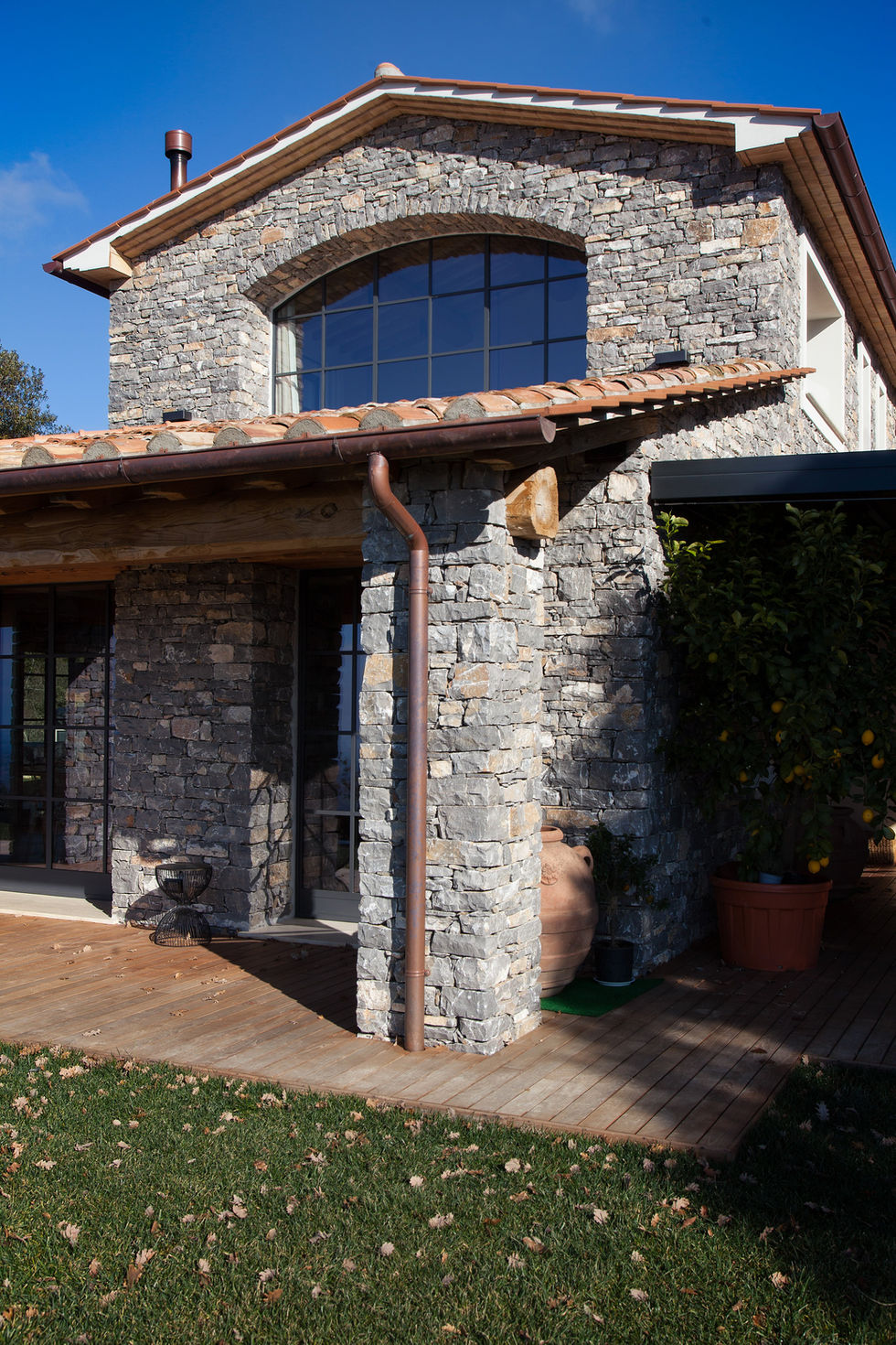 Exterior stone facade detail of Villa Bel Colle in Scansano, Maremma, showing dry-stacked local stone, terracotta roof tiles, copper downpipe, and terracotta amphora