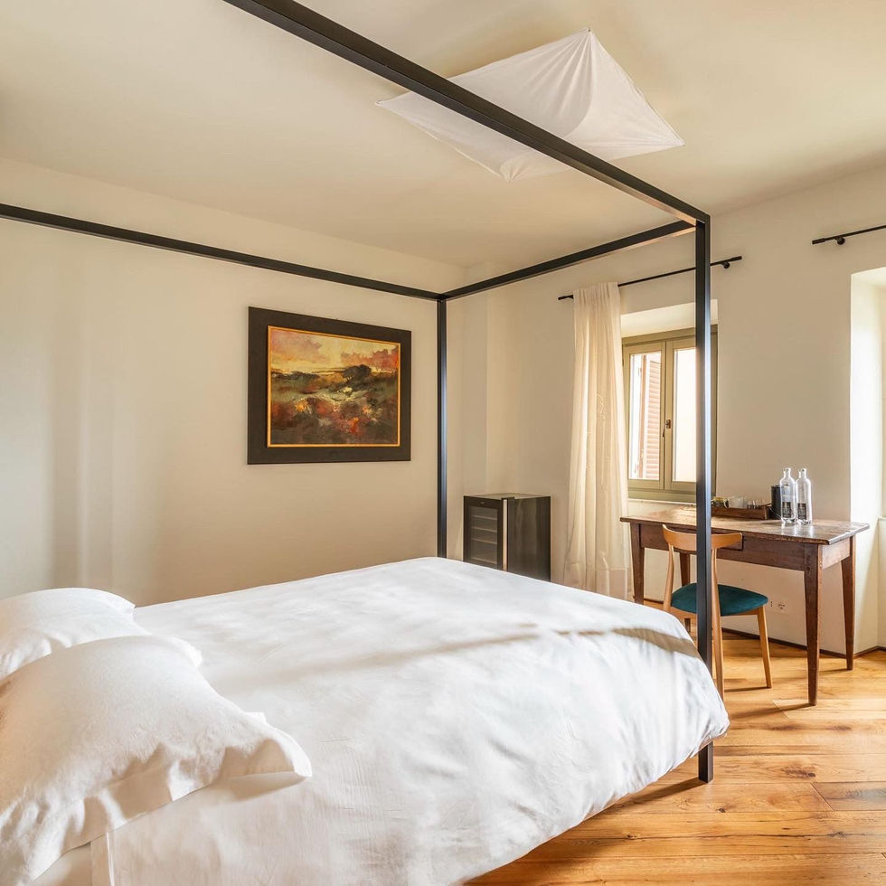Bedroom at Osteria Foro Civitella in Val di Chiana with black steel four-poster bed frame, white linen, oak parquet floor, landscape painting, and wooden writing desk by shuttered window