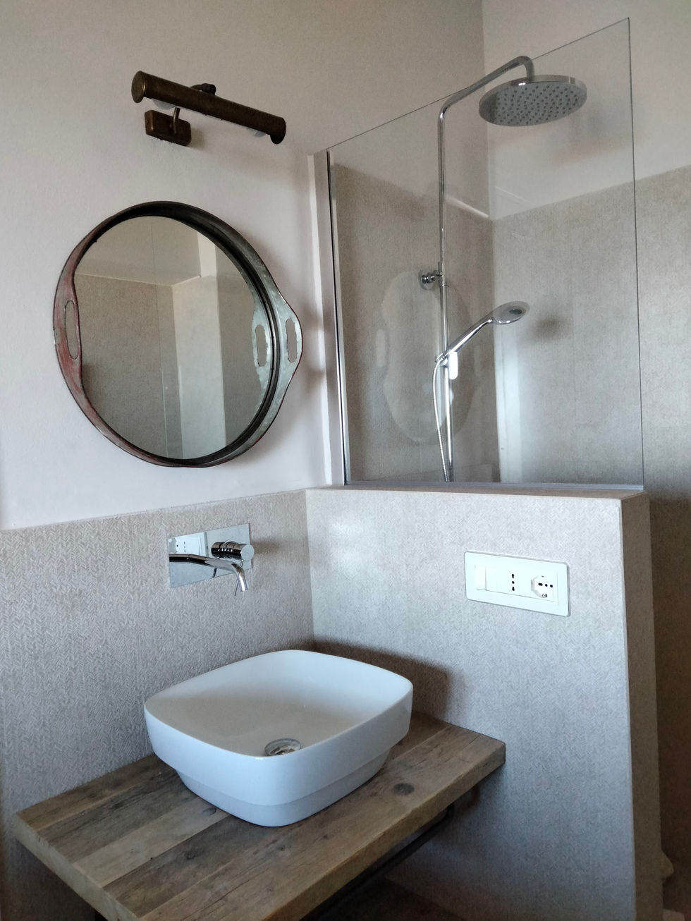 Compact bathroom of Villa San Feliciano with textured microcement walls, round mirror, wall-mounted chrome faucet, and glass shower enclosure, Magione, Umbria