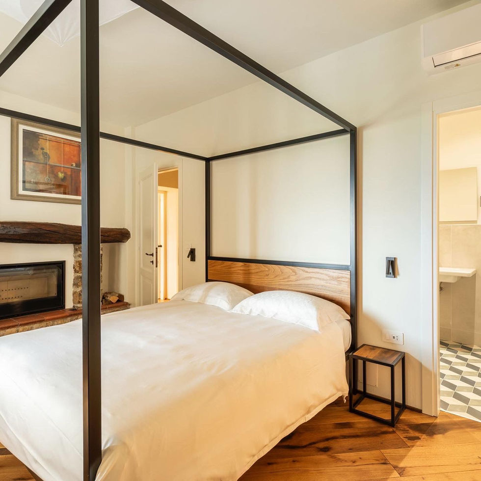 Guest bedroom at Osteria Foro, Civitella in Val di Chiana, with black steel four-poster bed, oak headboard, stone fireplace, and geometric-tile ensuite glimpse