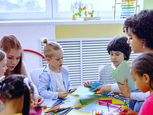 Children and a teacher gather around a table, making colorful paper crafts in a bright classroom.