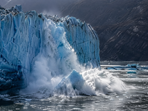 Massive glacier calving into the ocean as a large wall of ice collapses, sending ice chunks and spray into the water beneath steep icy cliffs.