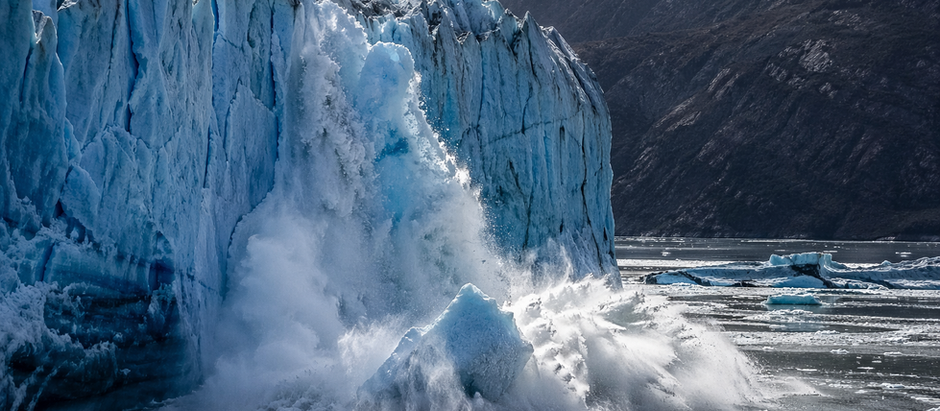 Massive glacier calving into the ocean as a large wall of ice collapses, sending ice chunks and spray into the water beneath steep icy cliffs.