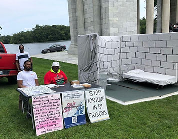 Three people sit behind a table with signs that say, "Stop Torture in RI prisons" and other messages. A mock solitary confinement cell is next to them.