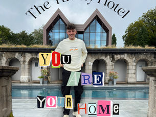 A person stands in front of a modern building with a pool, framed by stone arches and greenery. Above the person, curved text reads “The Tawny Hotel.” Overlaid on the image in colorful, cut-out letters is the phrase: “YOU ARE YOUR HOME.” The person is dressed casually in a light sweatshirt and dark pants, with white sneakers. The setting appears tranquil and inviting, blending contemporary architecture with natural surroundings.