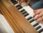 Hands playing a wooden piano keyboard with sheet music visible. The black and white keys contrast with the warm wood grain, creating a focused mood. Hospice volunteer
