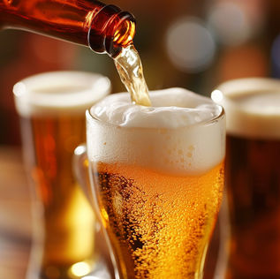 A bottle of beer is pouring into a tall, slender beer glass. The beer in the glass is a light amber color, and the poured beer forms a head of white foam. 