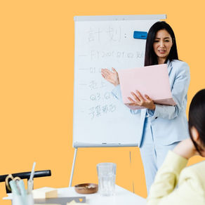 Woman in a light blue suit presents with a pink laptop by a whiteboard with text, facing a seated person. Background is pale orange.