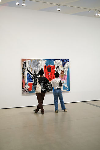The image shows two individuals standing in a modern art gallery, looking at a large, colorful abstract painting mounted on a white wall. The artwork features a mix of bold red, blue, and black colors, and incorporates various abstract shapes and figures. The visitors are closely observing the piece, emphasizing a quiet, contemplative atmosphere typical in art museums. The gallery space is minimalistic with a clean and open design, drawing focus to the artwork.