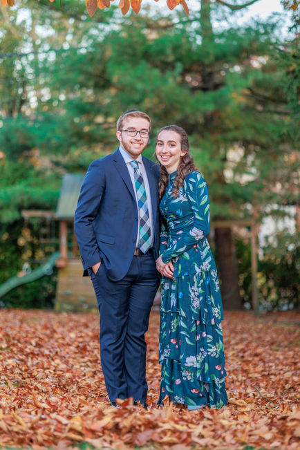 Engaged couple smiling in a wooded garden setting, the bride-to-be in a dark floral dress.