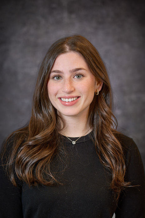 Woman with long brown hair in a black top, smiling confidently in a studio portrait.