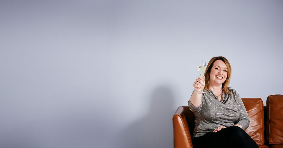 Smiling woman with shoulder-length blonde hair sitting on a brown leather sofa, raising a glass of prosecco in a toast. She is wearing a grey wrap-style top and black trousers, with a soft blue wall in the background — perfect brand image for celebrating success or relaxed confidence in business.