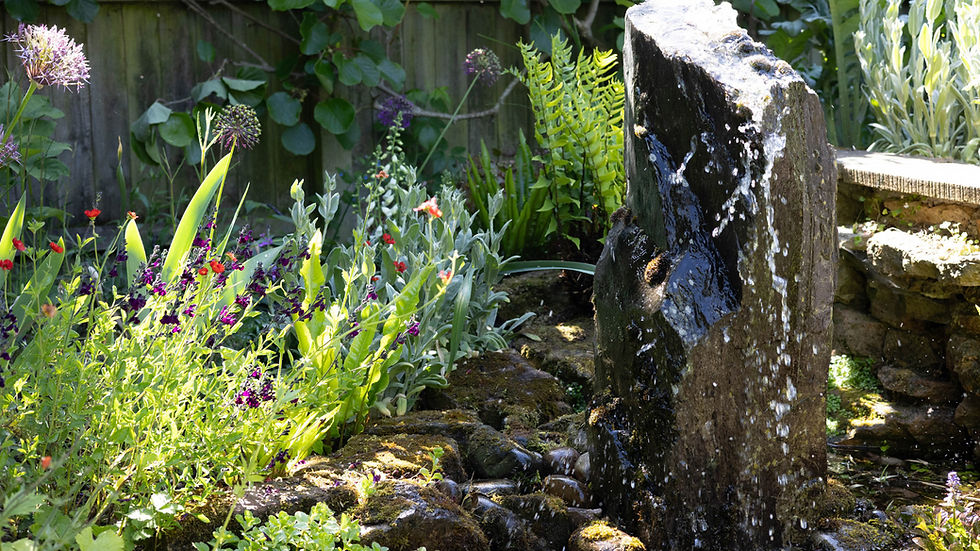A slate monolith with water bubbling out the top and cascading down onto a bed of cobbles. Surrounded by purple Salvias and green ferns.