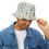 Thumbnail: Man wearing a white bucket hat with colorful doodle-style prints, styled with a denim jacket and white t-shirt, smiling sli