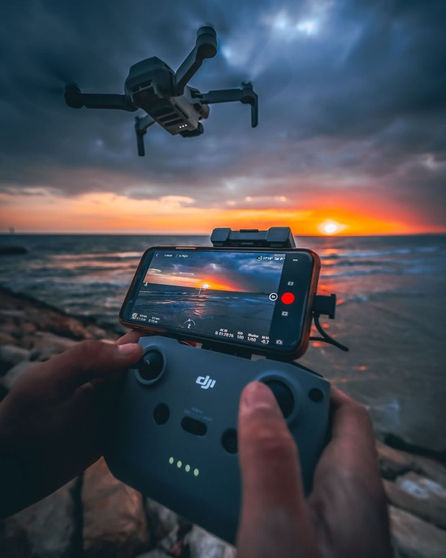 Mãos operam drone com pôr do sol e mar na tela.