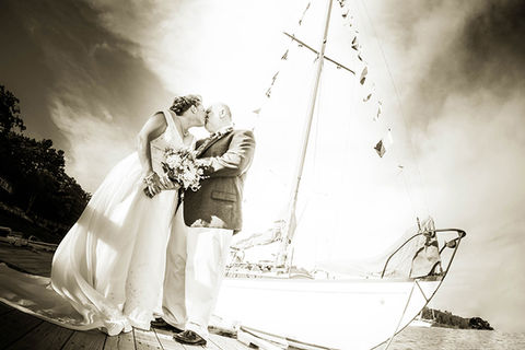 Black and white image.  Middle aged straight white couple kiss.  The bride wears wedding gown with long train and holds a large bouquet.  Groom is dressed in dark jacket and khaki pants.  Half the image is the bow of a stunning wooden yacht, dressed in flags.  The mast forms a cross that reaches up into a sky of clouds.  Boothbay Harbor, Maine