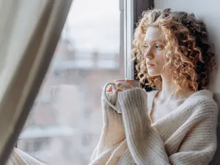 Femme assise près d’une fenêtre avec une tasse, pensive pendant un moment de repos.