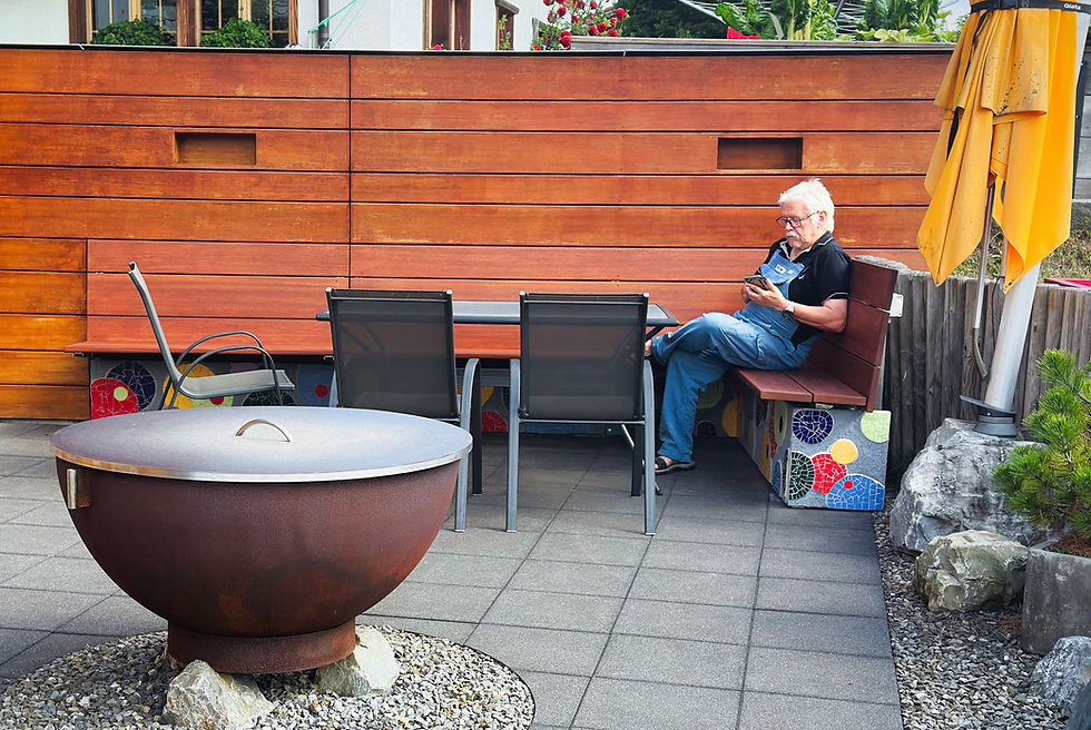 Fertig montierte Sitzbank mit bunter Mosaikverkleidung in Rot, Gelb, Blau und Grün an der Unterseite, auf einer Terrasse in Adelboden. Ein älterer Herr sitzt entspannt auf der Bank und schaut auf sein Handy, davor ein Gartentisch mit Stühlen und eine runde Feuerschale.