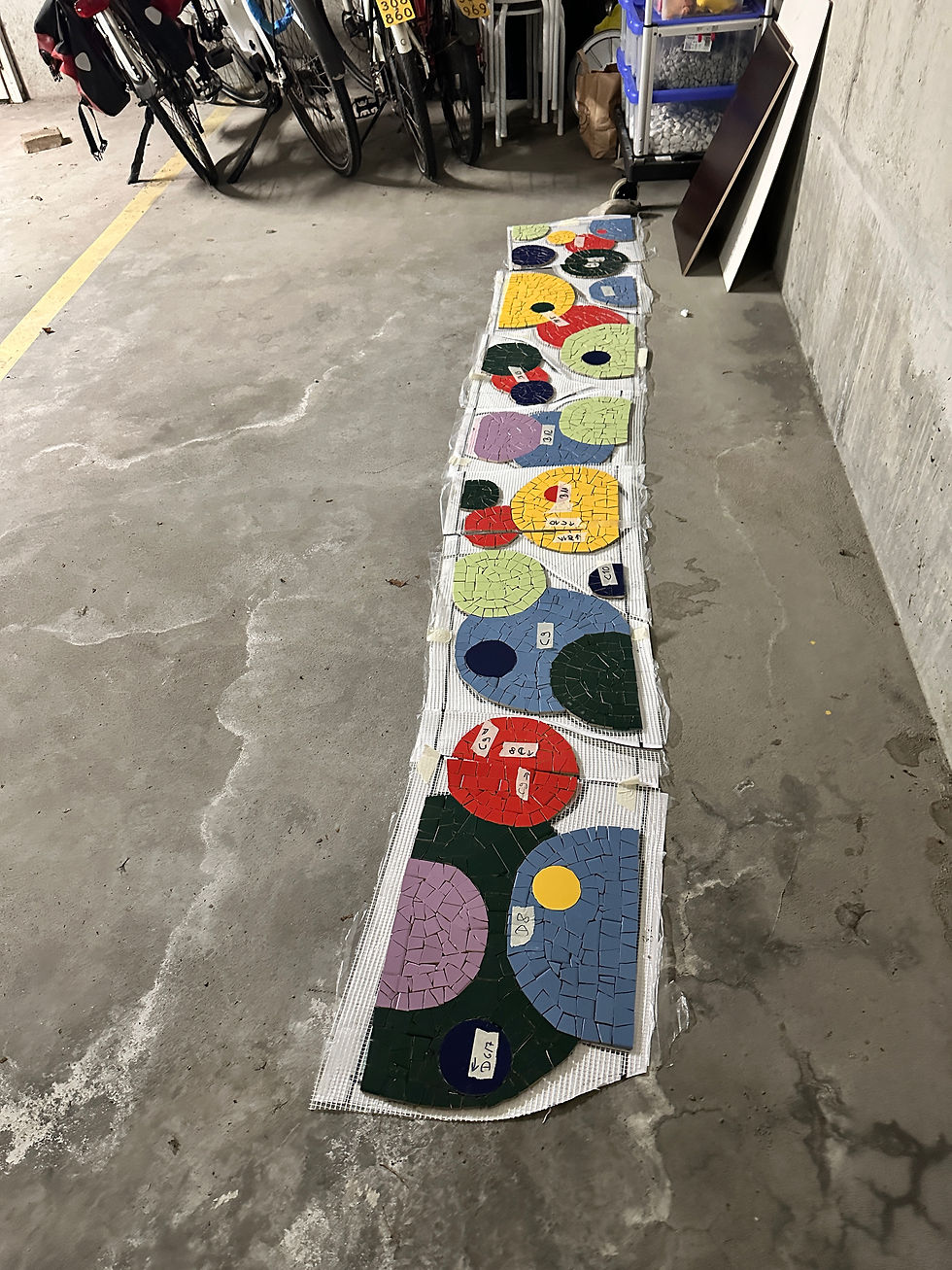 Ein langer, farbenfroher Streifen eines unfertigen Mosaik-Projekts liegt auf dem Betonboden einer Werkstatt. Die Mosaiksteine, in Formen von Kreisen und Halbkreisen in Gelb, Rot, Blau, Grün, Violett und Weiß, sind auf einem weißen Trägernetz ausgelegt. Viele der Mosaiksegmente sind mit handgeschriebenen Etiketten (z.B. 'C7', 'C8', 'D6') markiert, die auf einen Montageplan hindeuten. Im Hintergrund sind parkende Fahrräder und Lagerkisten auf rauer Betonoberfläche zu sehen.