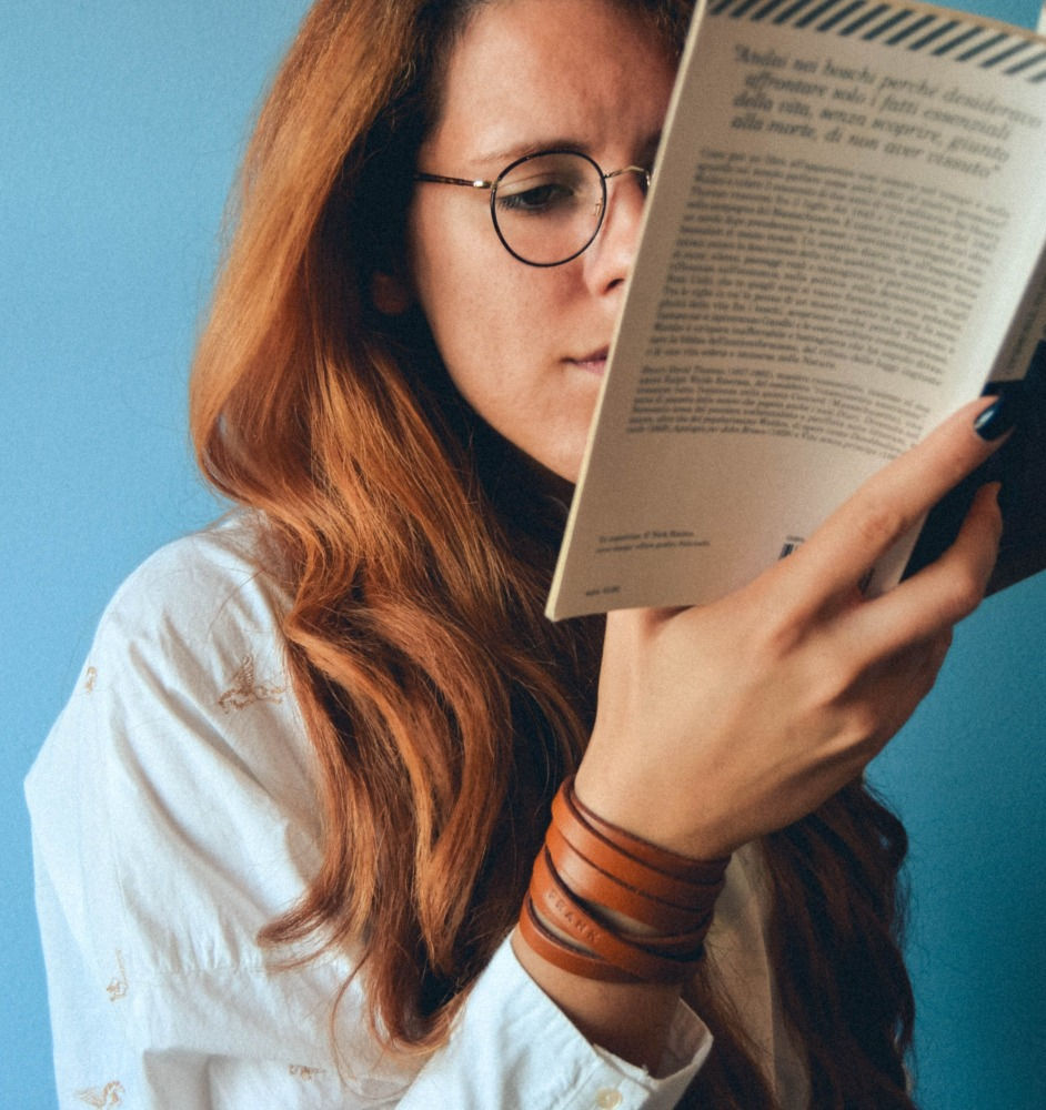 Parede ao fundo azul. Mulher ruiva usando óculos lê um livro que segura apenas com a mão direita. Ela veste camisa branca e uma pulseira de fita de couro.