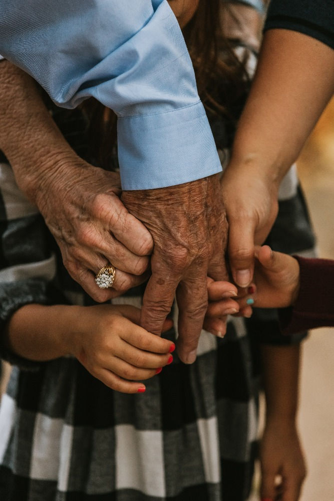 Mãos de diferentes idades, todas juntas. A mão que está ao centro é de um homem idoso. Ao redor dela, segurando quatro dos cinco dedos, estão uma mão de mulher idosa, duas mãos de crianças e uma mão de mulher adulta