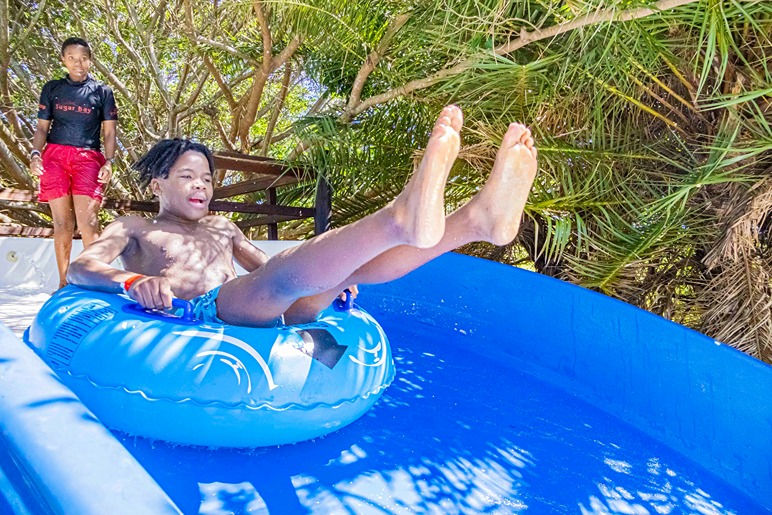 Shirtless man rides a blue inflatable in a pool slide while another man in a Sugar Boy shirt stands behind under palm trees. Sugar Bay Camp, Zinkwazi Beach, South Africa.