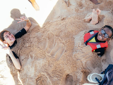 Two smiling people buried in beach sand with hands raised; one wears glasses and a red life vest. Sugar Bay Camp, Zinkwazi Beach, South Africa.