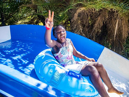 Smiling girl in a blue inflatable raft on a water slide, flashing a peace sign under palm trees. Sugar Bay camp zinkwazi beach south africa
