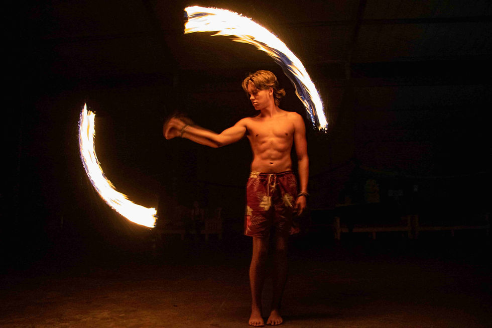 A shirtless Sugar Bay counselor spins flaming poi indoors, creating arcs of light against a dark background. Wearing patterned shorts, mood is intense.