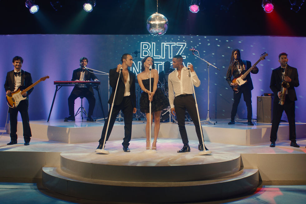 Band performing on stage with microphones, guitar, keyboard, and saxophone. Disco ball above, colorful lights. Text reads "BLITZ NATION". Energetic mood.