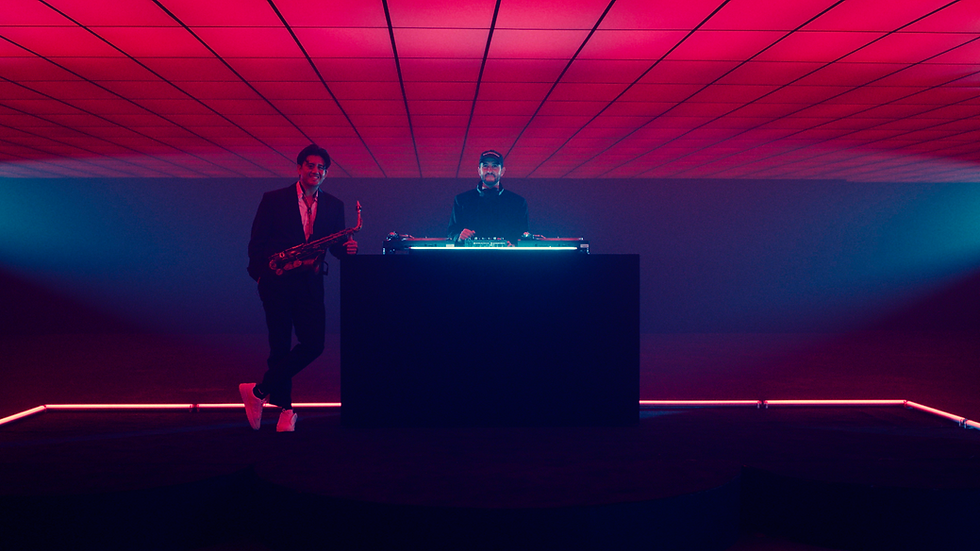Two musicians in a neon-lit room; one holds a saxophone, the other stands at a turntable. Red and blue lights create a vibrant atmosphere.