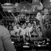 People in party attire cheer and dance under string lights with a DJ in the foreground, creating a lively and festive atmosphere.