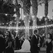 Wedding reception under string lights with guests in formal attire, palm trees in the background, creating a festive and elegant atmosphere.