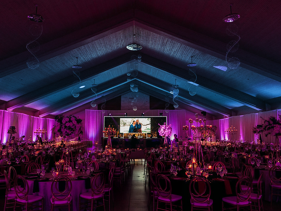 Elegant banquet hall with round tables, floral centerpieces, and purple up lighting. A large screen displays a photo slideshow.