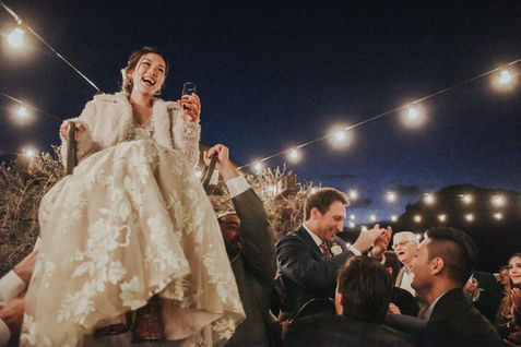 Bride lifted up on a chair during a lively outdoor wedding reception with string lights.