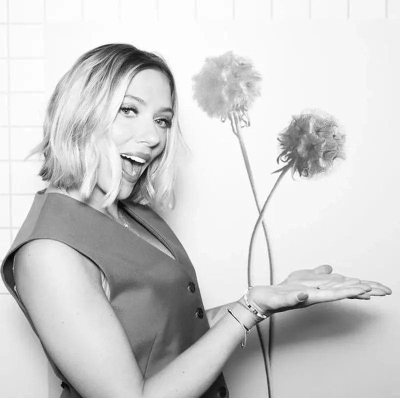 Smiling woman Scarlett Johansson in sleeveless dress gestures towards a drawing of two flowers on a white, checkered background. Black and white photo booth.