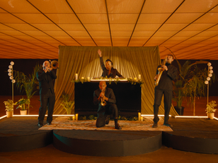 dj and live musician on stage with brass instruments, DJ behind a console, raising a hand. Orange ceiling, plants, and warm lighting create an energetic vibe.