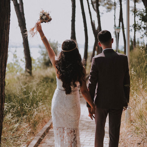 A bride in a lace gown joyfully raises her bouquet into the air, walking down a forest path with her partner in front of her, who is seen from the back in a formal suit.