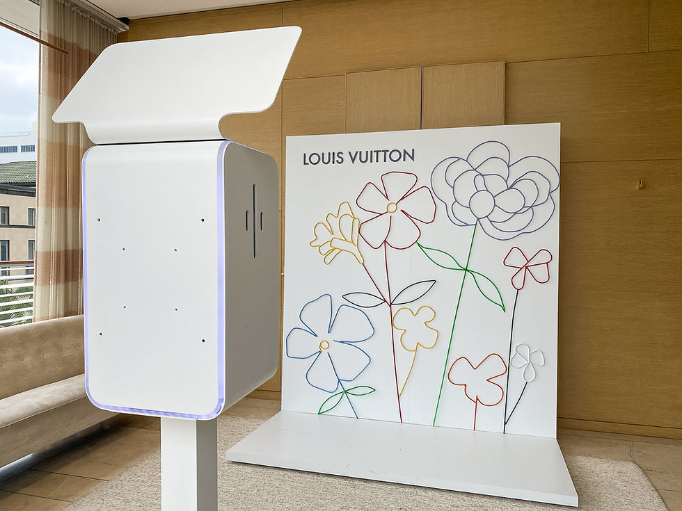 White photo booth next to a Louis Vuitton brand integration backdrop with colorful line art flowers. Indoor setting with neutral tones and soft lighting.