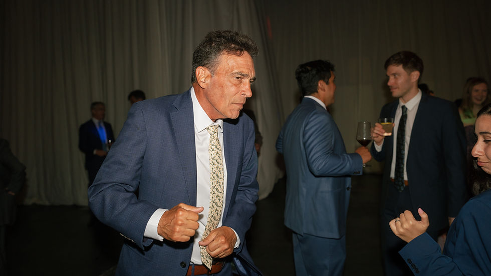 Older man in blue suit dancing energetically at a wedding event; others in background holding drinks, engaging socially. Mood is lively and upscale.
