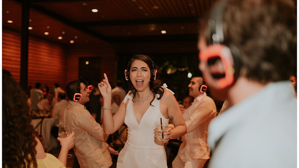 People wearing red silent disco headphones dance joyfully at a silent disco in a warmly lit room. A woman in white leads, holding a drink, smiling.