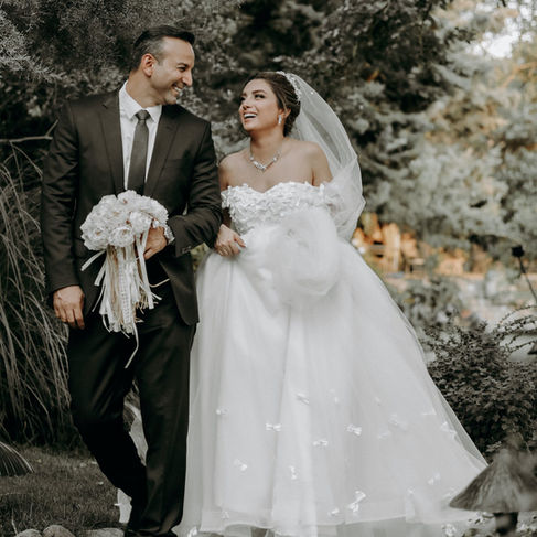 A beaming bride in a white ball gown and a groom in a black suit share a candid, joyful moment together, surrounded by lush greenery in an outdoor setting.