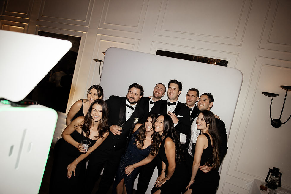 A group of people in formal attire pose and smile for a photo in front of a white backdrop, creating a joyful atmosphere at a wedding