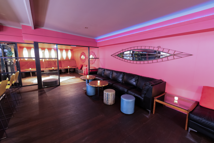 The Aberdeen Hotel creates a trendy lounge with pink walls, black leather sofas, blue and wood stools. Neon lighting and eye-shaped mirror create a cozy, modern vibe. 