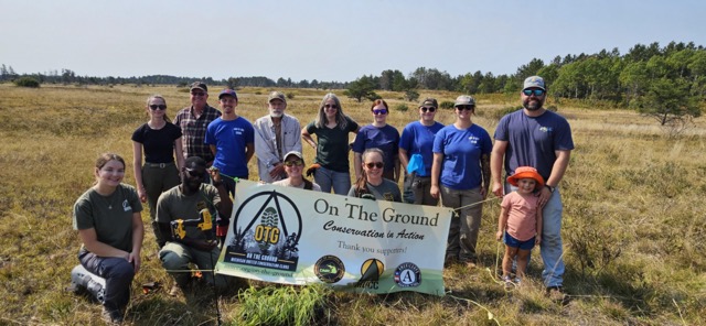 A dozen people pose for a photo with a sign with the group name "On The Ground"
