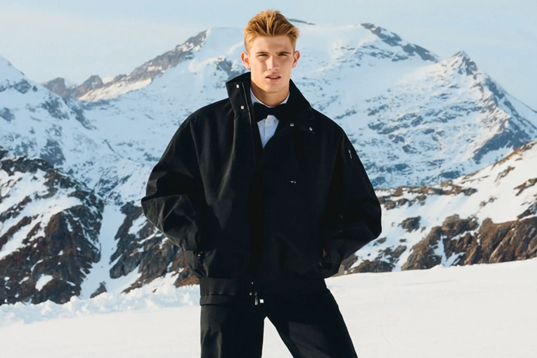 Male model in an oversized black Nike × Jacquemus ski jacket standing in front of snow-covered mountains during the Après Ski campaign.