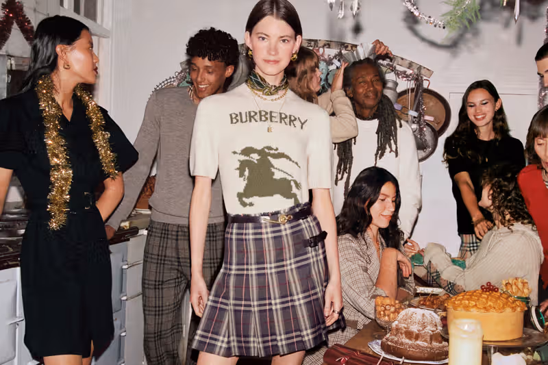 Models in Burberry holiday outfits standing around a festive dinner table, featuring the brand’s check patterns and seasonal styling.
