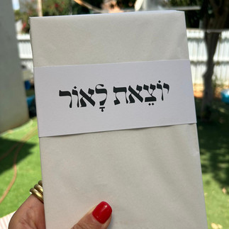 יציאת לאור text on a white paper. Hand holding the paper outdoors.