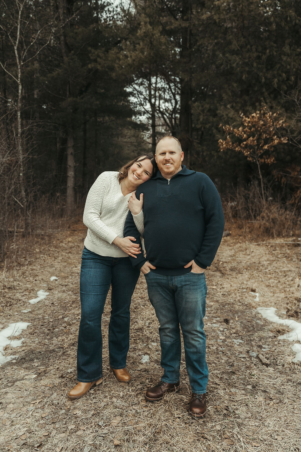 Outdoor engagement photo of couple standing on a trail with the woman's head on the man's shoulder and her arms wrapped around his. Photographed near Elk River, Minnesota.
