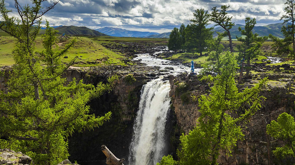 Waterfall cascades amid lush greenery under a cloudy sky. Trees and rocks surround the scene, creating a serene and natural landscape.